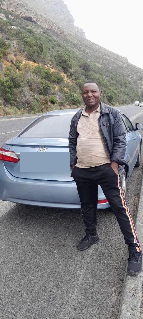 Fanuel Chinovunye standing beside his light blue car, with the mountain behind him